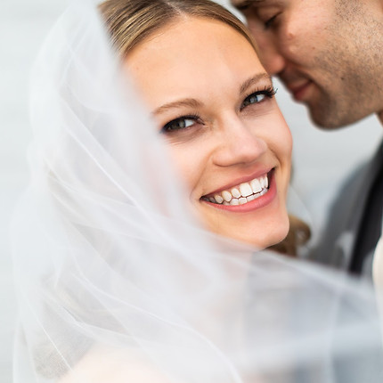a beautiful wedding photo of the bride glancing at the camera through her veil with her groom behind her